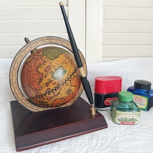 Op de afbeelding: Een bureau-set met een globe in vintage-stijl op een donkerbruine houten voet met een zwarte en gouden vulpen. Naast de voet staan drie glazen inktpotjes met rode, groene en blauwe doppen en etiketten, waaronder een flesje van J. Herbin.