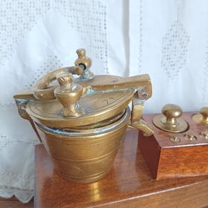 May include: Antique brass apothecary scale with a small pot and lid, and a wooden box with weights. The scale has a handle and a hinged lid. The wooden box has a polished finish. The background is a white lace curtain.