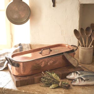 Puede incluir: Una pescadera ovalada de cobre martillado con tapa a juego y dos asas laterales. El utensilio de cocina está sobre una tabla de cortar de madera junto a dos pescados crudos enteros, una espátula de metal y un manojo de hierbas frescas atadas.