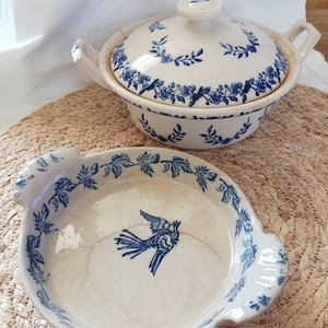 Puede incluir: Una fuente de cerámica color crema con tapa y un cuenco poco profundo a juego, ambos con un patrón de enredadera floral azul. El cuenco muestra una ilustración de un pájaro azul en el centro y la fuente tiene dos asas laterales.