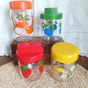 Könnte beinhalten: Vier Vintage-Gläser mit farbigen Deckeln und Fruchtdesigns. Die Gläser haben orange, grüne, rote und gelbe Deckel. Die Fruchtdesigns umfassen Orangen, Pflaumen, Erdbeeren und Zitronen. Ideal für die Küchenaufbewahrung.