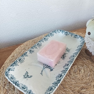 May include: A rectangular, off-white ceramic soap dish with a blue floral and butterfly design. A pink bar of soap rests in the center. The dish is placed on a woven mat.
