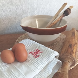 Op de afbeelding: Een crème en bruine keramische mengkom met houten lepels, een deegroller en een witte wafelhanddoek geborduurd met een rode initiaal. Drie bruine eieren liggen op de handdoek, wat wijst op een bakthema.