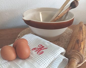 May include: A cream-coloured ceramic mixing bowl with a brown rim, two wooden spoons, a wooden rolling pin, three brown eggs, and a white waffle-weave kitchen towel featuring a red embroidered letter M monogram, all arranged for baking preparation.