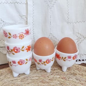 May include: White ceramic egg cups with a floral design in pink and orange. The egg cups are stacked and also holding brown eggs. The egg cups have three small feet.