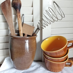 Peut inclure: Ensemble d'ustensiles de cuisine et de vaisselle vintage. Un pot en céramique marron contient des cuillères en bois, une spatule et une planche à beurre. Un fouet en métal et trois tasses marron à intérieur jaune sont empilés.