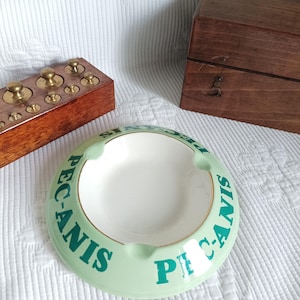 May include: A round, light green ceramic ashtray with a white interior and a gold-toned rim. The exterior features the dark green text "PEC-ANIS" printed repeatedly around the circumference. The ashtray has three notches for resting items.