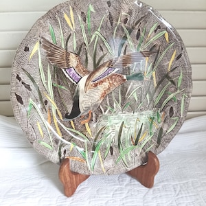 May include: A round decorative plate featuring a detailed illustration of a mallard duck in flight over a marshy pond. The background displays a textured brown cross-hatch pattern with green reeds and cattails. The duck has a green head and purple wing patches.
