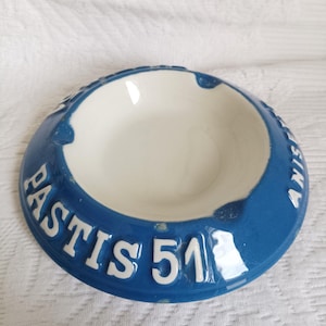 Könnte beinhalten: Ein runder Aschenbecher aus Keramik mit weißer Mitte und breitem blauem Rand. Auf dem Rand befinden sich erhabene weiße Buchstaben mit der Aufschrift PASTIS 51 und ANISE. Der Aschenbecher hat drei Einkerbungen am Rand und eine glänzende Oberfläche.