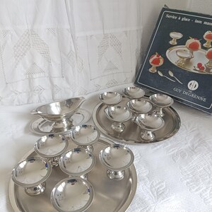 Peut inclure: Un ensemble de plats et de plateaux de service argentés, comprenant de petits bols à piédestal, une saucière et des plateaux de service. L'ensemble est disposé sur une surface blanche et texturée, avec une boîte portant l'inscription "Service à glace".
