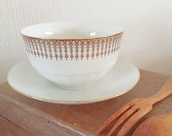 May include: A white porcelain bowl and matching saucer featuring a gold-colored decorative band with a repeating geometric pattern along the rim. The bowl has a subtle ribbed texture on the lower exterior, and both pieces are finished with gold trim.
