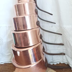 May include: A stack of six copper cooking pots with dark metal handles. The pots are different sizes, with a shiny, reflective surface. The handles curve upwards. The pots are stacked on a woven mat.