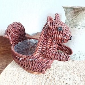 May include: A brown wicker squirrel-shaped basket with a clear plastic liner. The basket has a detailed woven texture, with a tail, ears, and a small black button for an eye. The basket is sitting on a woven mat.