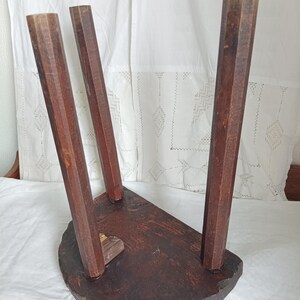 May include: A small, antique wooden stool with three angled legs. The stool has a dark brown, semi-circular seat and is made of solid wood. The legs are faceted and taper towards the base, showcasing the natural wood grain.