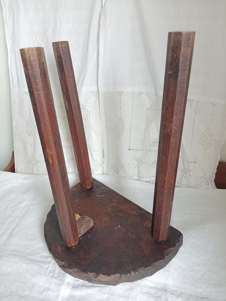 May include: A small, antique wooden stool with three angled legs. The stool has a dark brown, semi-circular seat and is made of solid wood. The legs are faceted and taper towards the base, showcasing the natural wood grain.