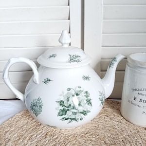 May include: White ceramic teapot with a lid, featuring a green floral pattern. The teapot has a curved handle and spout. A white ceramic jar with text is visible to the right. The teapot sits on a woven mat.