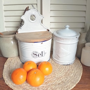 Pode incluir: Natureza morta com utensílios de cozinha vintage e laranjas frescas. Saleiros e potes de especiarias esmaltados brancos com tampas sobre um tapete tecido. O saleiro tem a palavra "Sel". Quatro tangerinas laranja brilhantes em primeiro plano.