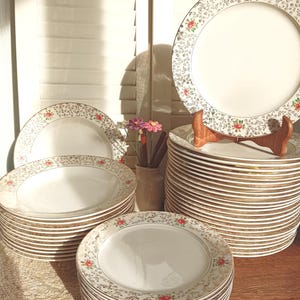 Peut inclure: Ensemble d'assiettes en céramique blanche avec un motif floral et un bord doré. Plusieurs piles d'assiettes sont visibles, certaines étant présentées sur un présentoir en bois. Le motif floral comprend des roses rouges et de petites feuilles vertes.
