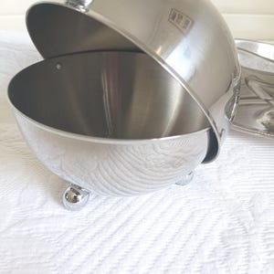 May include: A polished stainless steel serving bowl with a spherical, hinged lid that opens to reveal a deep interior. The bowl features a smooth, reflective finish and is supported by three small, rounded ball-shaped feet at the base.