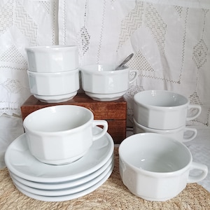 May include: A collection of white ceramic teacups and saucers. The cups have a unique octagonal shape and small handles. The saucers are round and stackable. The set includes several cups and saucers, perfect for serving tea or coffee.