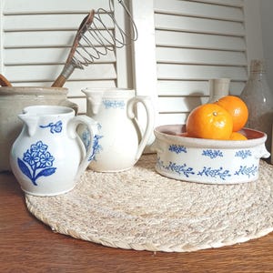 Può includere: Una collezione di stoviglie da cucina in ceramica bianca vintage con disegni floreali blu. Include brocche, una pirofila con arance e una frusta. Gli oggetti sono disposti su un sottobicchiere intrecciato, creando un'estetica rustica e country.