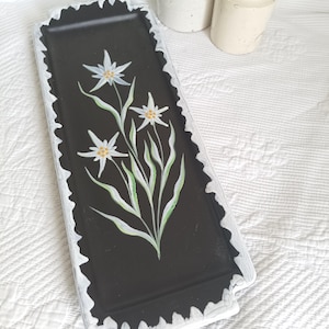 May include: A rectangular black serving tray featuring a hand-painted edelweiss flower design with white petals and yellow centers. The tray has a scalloped, distressed white border along the edges, providing a contrast to the dark center.