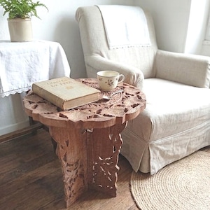 May include: A round, brown wooden side table with intricate floral carvings on the top and base. The table features a folding design with three interlocking legs. A beige book titled Contes de Fees and a small ceramic teacup sit on the surface.