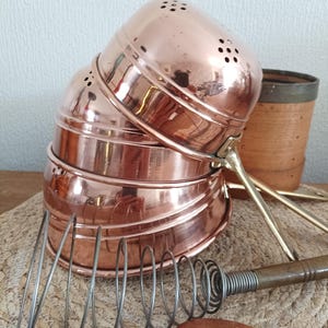 Op de afbeelding: Een stapel van drie gepolijste koperen vergieten met geperforeerde bovenkanten en messing handgrepen, vergezeld van een metalen spiraalgarde met houten handvat en een houten lepel. De koperen oppervlakken zijn reflecterend en warm oranje.