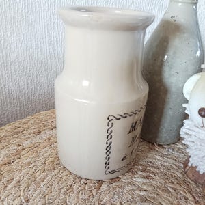 May include: A cream-coloured ceramic pitcher with a wide rim and a cylindrical body. The front features a dark brown decorative border enclosing text that reads MAISON, Miel, &agrave; l'Ancienne. The pitcher has a smooth, glossy finish.