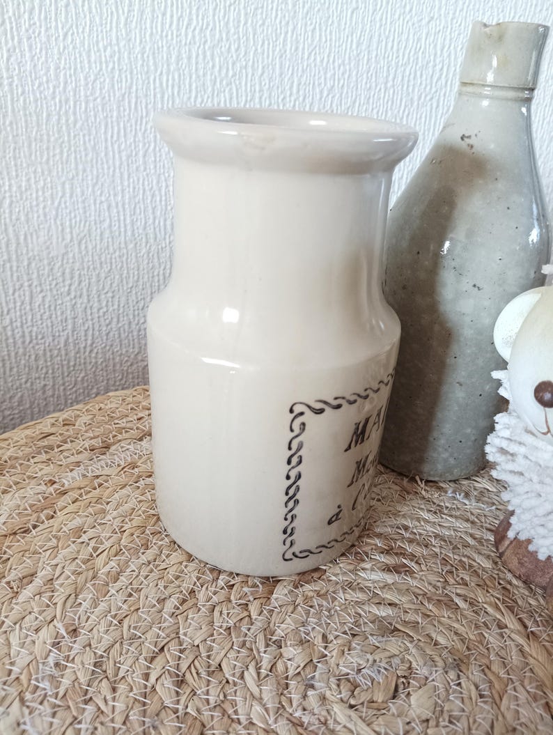 May include: A cream-coloured ceramic pitcher with a wide rim and a cylindrical body. The front features a dark brown decorative border enclosing text that reads MAISON, Miel, &agrave; l'Ancienne. The pitcher has a smooth, glossy finish.