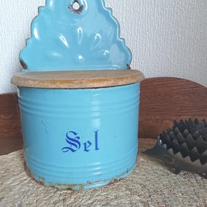 Op de afbeelding: Een vintage, lichtblauwe emaille zoutpot met een houten deksel en decoratieve achterplaat. Het woord "Sel" is in blauw schrift op de voorkant gedrukt. De doos heeft een cilindrische vorm en is waarschijnlijk voor gebruik in de keuken.