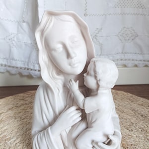 Puede incluir: Una escultura de cerámica blanca de una mujer sosteniendo a un niño. La mujer lleva un velo y sostiene al niño cerca. El niño extiende la mano hacia la mujer. La escultura está sobre una estera tejida.