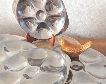 May include: A set of three circular stainless steel oyster plates, each featuring six individual shell-shaped indentations and a central circular well, accompanied by a small, brown, glazed ceramic bird figurine.