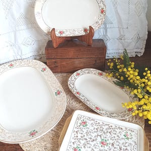 May include: A collection of vintage serving platters and a tray. The platters are white with a gold floral border and red rose accents. One platter is displayed on a wooden stand. The tray has a floral pattern.