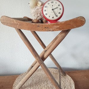 Peut inclure: Un tabouret pliant en bois vieilli supportant un réveil rouge et un mouton en peluche. L'horloge a un cadran blanc avec des chiffres noirs. Le mouton est blanc avec un visage beige. Le tabouret est en bois brun clair.
