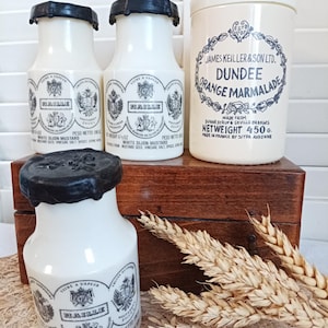 Puede incluir: Un conjunto de envases de alimentos vintage. Tres botellas blancas de mostaza Dijon blanca "HAILLE" con tapas y etiquetas negras. También hay un tarro color crema de "DUNDEE ORANGE MARMALADE", junto con espigas de trigo y una caja de madera.