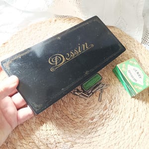 May include: A rectangular black metal box with the word Dessin printed in gold script on the lid. Beside the box are several silver metal dip pen nibs and a small green rectangular paper box labeled Henry Superieure Blanzy Conte Gilbert.