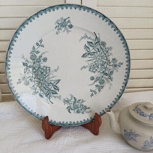 May include: A round white ceramic plate featuring a dark teal floral pattern and a decorative border. The plate is displayed on a wooden stand next to a small white teapot with a blue floral design, resting on a white textured fabric surface.
