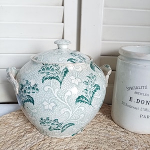 Puede incluir: Un cuenco redondo con tapa y diseño floral verde y blanco. El cuenco tiene dos asas y está colocado sobre una esterilla tejida. A la derecha hay un tarro de cerámica blanca con texto.