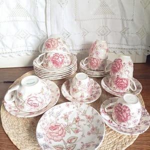 Könnte beinhalten: Eine Sammlung von Vintage-Tassen und Untertassen mit einem floralen Rosenmuster in Rosa und Grün. Die Tassen sind gestapelt, und die Untertassen sind auf einer gewebten Matte angeordnet. Der Hintergrund zeigt einen weißen Spitzenvorhang.