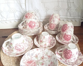 May include: A collection of white ceramic teacups and saucers featuring a pink and green floral pattern. The set includes several stacked cups and saucers, along with individual pieces arranged on a woven mat, showcasing detailed rose motifs.