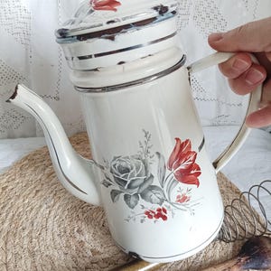 May include: A cream-coloured enamel coffee pot featuring a floral design with a grey rose and red tulips. The pot has a matching lid, a curved spout, and a side handle. There is visible wear and chipping along the rim and lid edges.