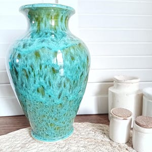 Peut inclure: Un grand vase en céramique turquoise avec une finition brillante. Le vase a un corps large et un bord évasé. La couleur turquoise se mélange à des nuances de vert, créant un effet marbré. Petits pots en céramique blanche avec couvercles en liège en arrière-plan.