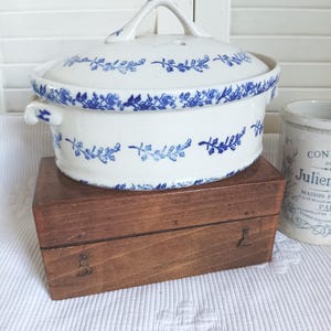May include: An oval ceramic casserole dish with a lid, featuring a white base and a blue floral vine pattern. The dish has two small side handles and a central lid handle. It is positioned on a rectangular wooden box with metal hinges.
