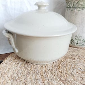 Puede incluir: Cazuela de cerámica blanquecina con tapa, asas laterales y borde decorativo. La tapa presenta un pequeño pomo texturizado. Un jarrón de estilo vintage con motivos florales verdes es parcialmente visible en el fondo.