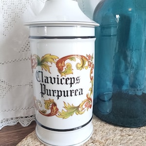 Peut inclure: Un pot d'apothicaire en céramique blanche avec couvercle assorti, doté d'un liseré noir et d'une illustration botanique décorative dans des tons orange, jaune et vert. Le texte Claviceps Purpurea est imprimé en police gothique noire sur le devant.