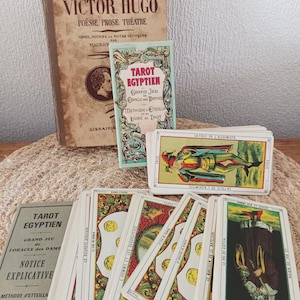 Puede incluir: Un conjunto de cartas de tarot antiguas y libros relacionados. El libro principal se titula "L'Oeuvre de Victor Hugo". Las cartas presentan ilustraciones detalladas y texto en francés. También hay un libro más pequeño, "Tarot Egyptien".