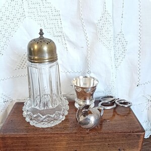 Peut inclure: Un salière en verre transparent avec un couvercle en métal argenté et une base décorative. À côté, une petite tasse argentée, un petit pichet argenté et trois anneaux argentés. Les objets sont sur une boîte en bois.