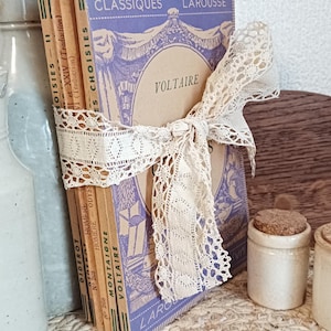 Peut inclure: Une pile de livres anciens attachés avec un ruban de dentelle crème. Les livres ont des couvertures bleues et beiges avec du texte français, dont "VOLTAIRE" et "CLASSIQUES LAROUSSE". Les dos montrent les noms d'auteurs et les numéros de livres.