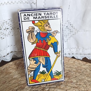 Peut inclure: Boîte de cartes de tarot vintage avec le texte "ANCIEN TAROT DE MARSEILLE". La boîte présente une illustration colorée d'une figure en bleu, rouge, jaune et or, portant un sac et un bâton. La boîte est rectangulaire.